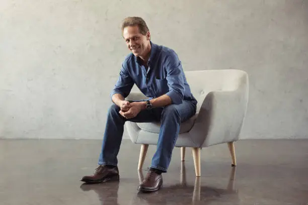 Photo of casual middle aged man smiling while sitting on armchair indoors