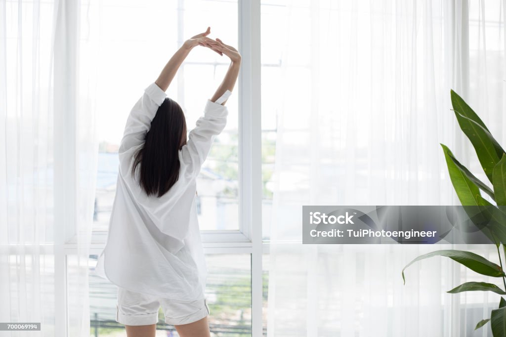 Asian woman waking up in her bed fully rested and open the curtains in the morning to get fresh air Asian woman waking up in her bed fully rested and open the curtains in the morning to get fresh air. Morning Stock Photo