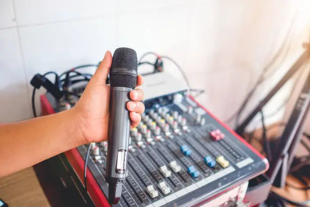 Photo of Hand holding wireless microphone against audio synthesizer electronic music instrument sound mixer machine in background