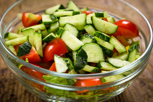 Salad with plenty of cut vegetables on glass bowl on rustic wooden table, closeup