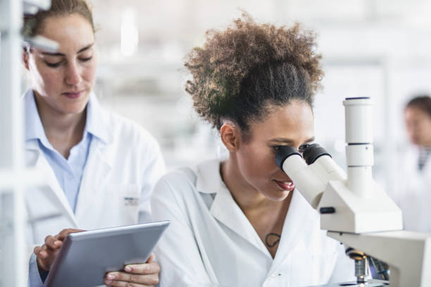 istock Female Scientist Looking Through A Microscope With Colleague Beside 700211792