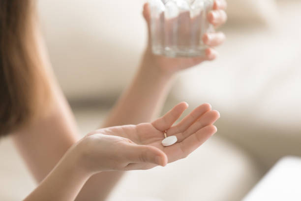 Close up photo of round white pill in female hand Close up photo of one round white pill in young female hand. Woman takes medicines with glass of water. Daily norm of vitamins, effective drugs, modern pharmacy for body and mental health concept painkiller stock pictures, royalty-free photos & images