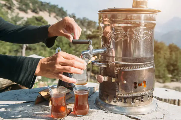 Drinking turkish tea outdoors. Hand holding spoon and tulip-shaped tea glass with traditional tea making pot Samovar