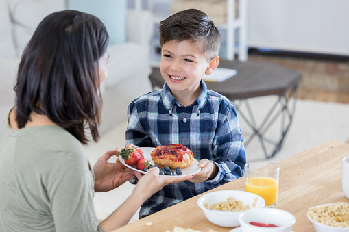 Cheerful young boy enjoys breakfast with his mom. She is handing him a plate of food. Bowls of cereal and orange juice are on the table.