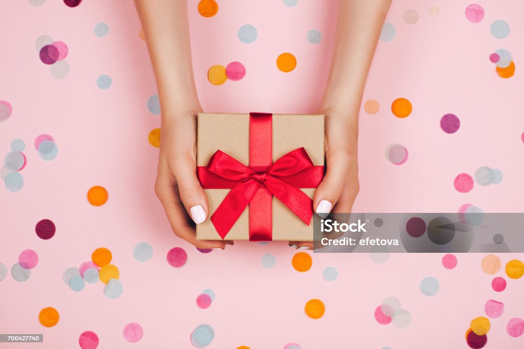 Hands holding present box Woman hands holding present box with red bow on pastel pink background with multicolored confetti. Flat lay style. Gift Stock Photo