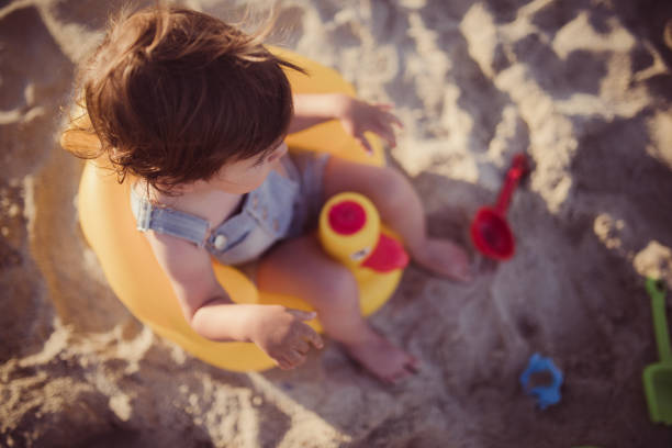 Child wearing inflatable armbands / water wings / swimmies / floaties, playing with sea sand, sitting on water's edge, wearing swimsuit and hat protect against the sun UV.