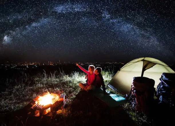 Photo of Night camping near the town. Young couple enjoying near campfire and tent, looking at beautiful night sky full of stars and enjoying night scene. Woman is pointing at the sky. Astrophotography