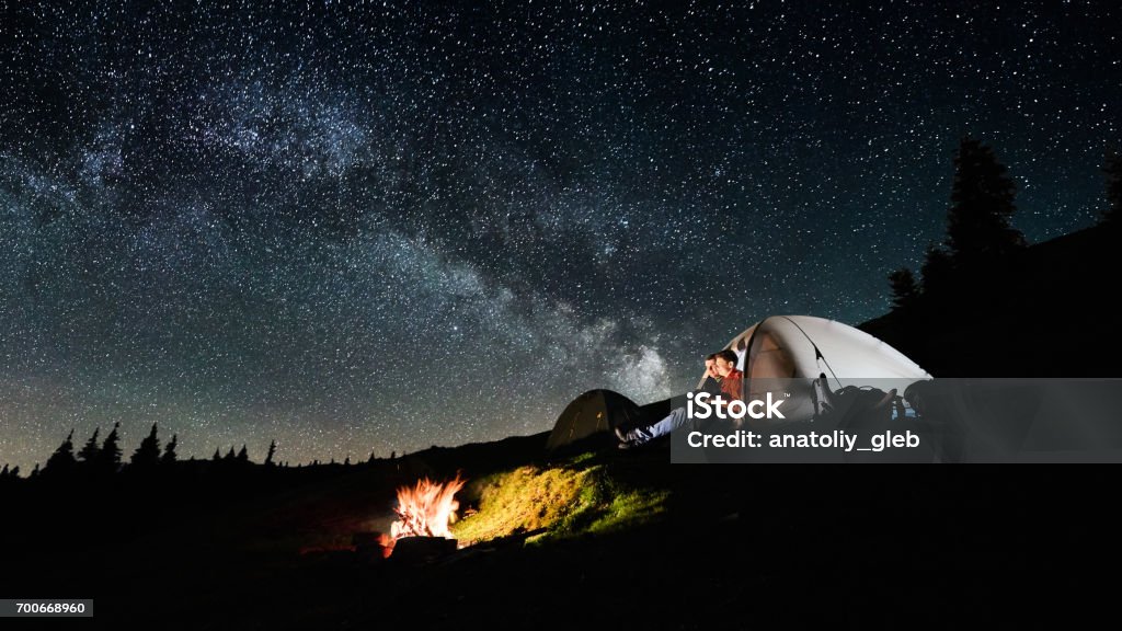Night camping. Man and woman tourists sitting in the illuminated tent near campfire under amazing night sky full of stars and milky way. Long exposure. Picture aspect ratio 16:9 Friendship Stock Photo