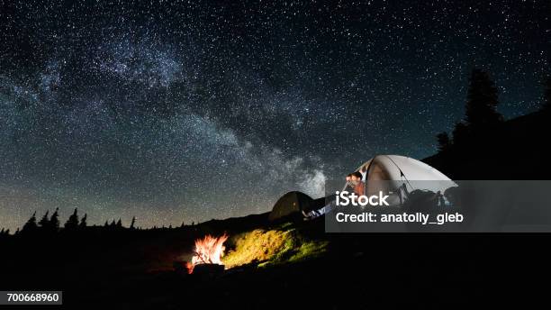 Night Camping Man And Woman Tourists Sitting In The Illuminated Tent Near Campfire Under Amazing Night Sky Full Of Stars And Milky Way Long Exposure Picture Aspect Ratio 169 Stock Photo - Download Image Now