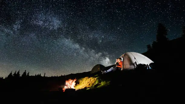 Night camping. Man and woman tourists sitting in the illuminated tent near campfire under amazing night sky full of stars and milky way. Long exposure. Picture aspect ratio 16:9