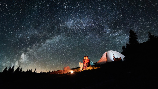 Noche de campamento. Los turistas de la romántica pareja tienen un descanso en una fogata cerca de la tienda iluminada bajo el increíble cielo nocturno lleno de estrellas y vía Láctea. Fotografía astronómica. Imagen relación de aspecto 16:9 photo