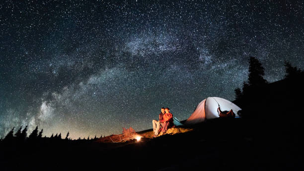 夜のキャンプ。ロマンチックなカップルの観光客は、素晴らしい夜満天の星と天の川の下で照らされたテント近くのキャンプファイヤーで残りを持っています。天体写真。アスペクト比 16:9 � - friendship camping night campfire ストックフォトと画像