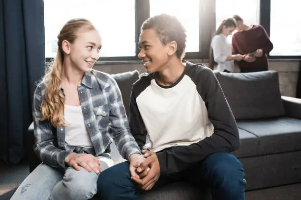 Photo of Happy teenage couple sitting on sofa and holding hands with friends standing behind, teenagers having fun concept