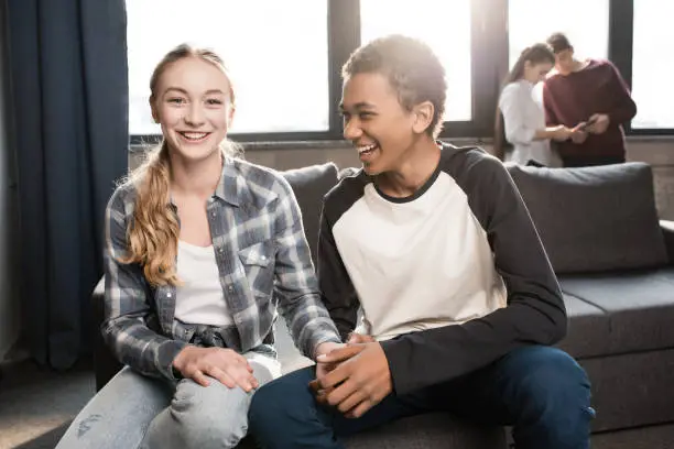 Photo of Happy teenage couple sitting on sofa and holding hands with friends standing behind, teenagers having fun concept