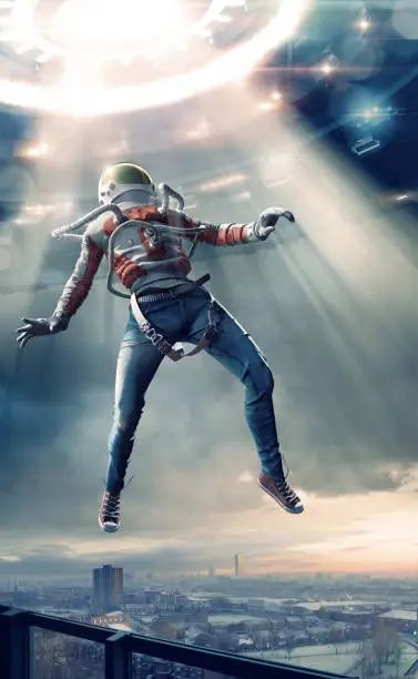 Conceptual image of a young man dressed in skinny jeans, sneakers, studded belt, spacesuit jacket and helmet levitating in mid air in a beam of light. The astronaut has moved from a balcony towards a UFO overhead with bright lights.  He could be an alien or being abducted from a city.