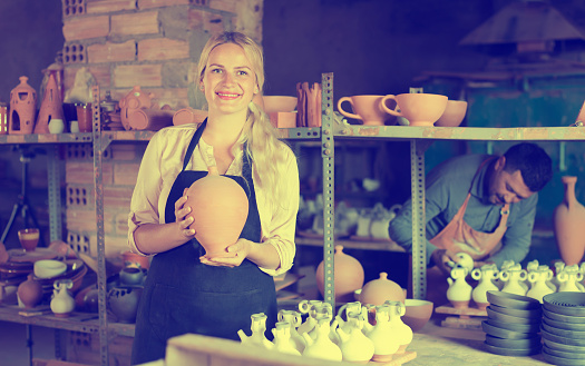 Laughing woman potter in apron carrying ceramic vessels in atelier