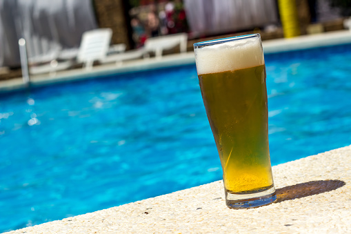 glass of cold beer sitting on edge of blue swimming pool