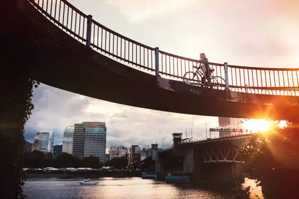 An adult woman walks her bicycle up a ramp from the waterfront Eastbank Esplanade in Portland, Oregon, connecting to the Burnside bridge.  The shine sets on the city in bright orange color.