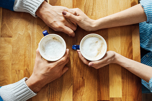 Directly above shot of young couple holding hands at cafe table. Cropped image of man and woman are with coffee cups on wood. They are spending leisure time in cafeteria.