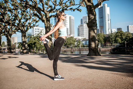 Young woman in sportswear having a morning exercise stretching in the park with skyscrapers on the background in Frankfurt city