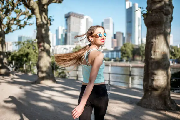 Young woman in sportswear walking and looking back in the park with skyscrapers on the background in Frankfurt city