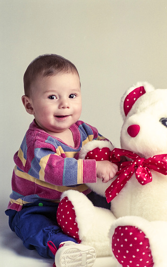 Baby boy playing with white (accented with red fabric with white hearts) large stuffed bear.  The stuffed bear is almost the same size as baby.