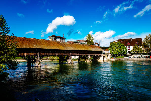 The Covered Swiss timber bridge was built in 1726 for pedestrians to cross the Aare river in Thun, Switzerland. The bridge was built as part of the upper (Scherzlig) river lock at the Aare in Thun. The wooden trestle bridge is covered and was built in 1726 for pedestrians to cross the Aare  (also called the Aar) river, a tributary of the High Rhine, flows next to an old building in the town of Thun, Switzerland. It is located in the Swiss alps and was shown in autumn. The river lock is also a site for river surfing.