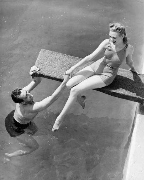 Woman sitting on diving board, man grasping her hand (B&W), elevated view  one piece swimsuit photos stock pictures, royalty-free photos & images