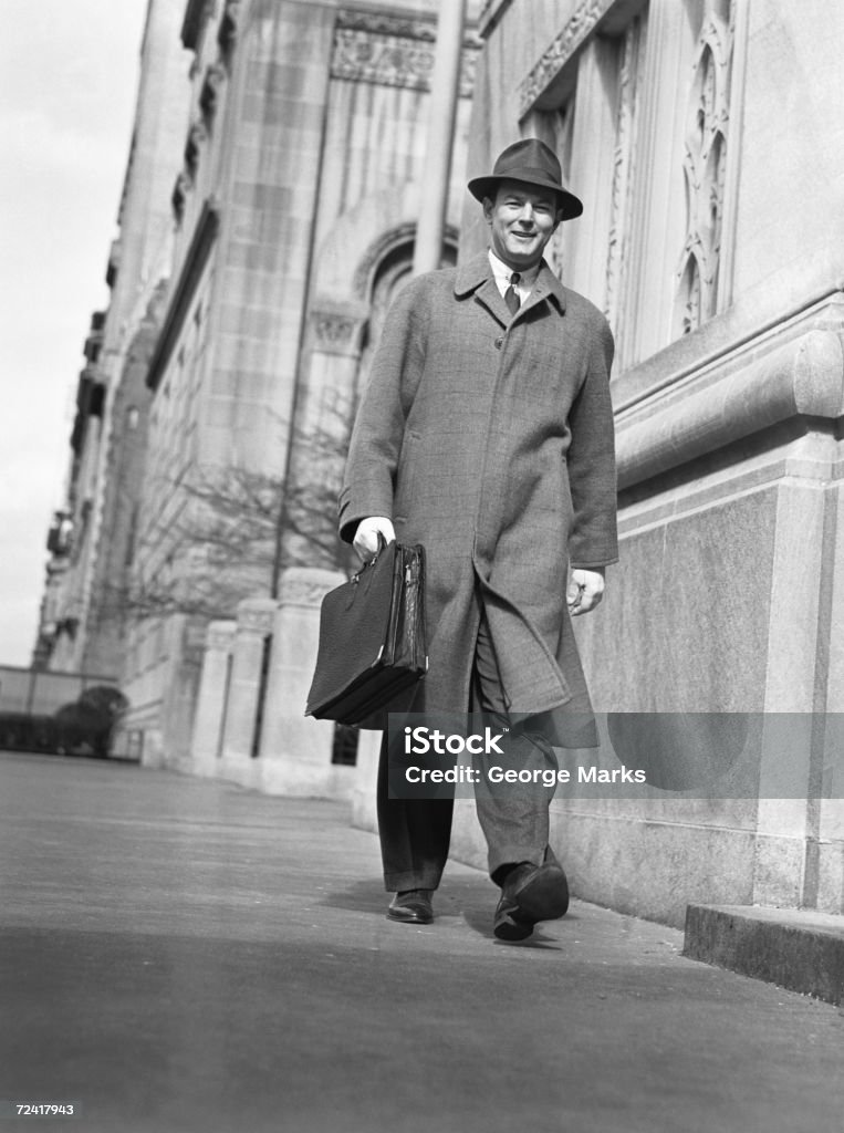 Man in overcoat and hat walking on sidewalk, (B&W), low angle view  1950-1959 Stock Photo