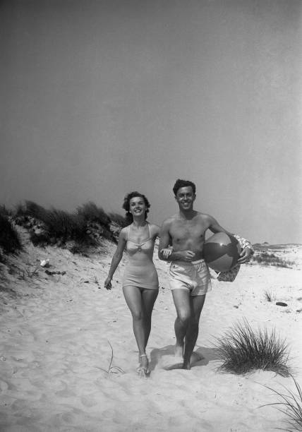 Couple walking on beach, man carrying ball, (B&W)  one piece swimsuit photos stock pictures, royalty-free photos & images