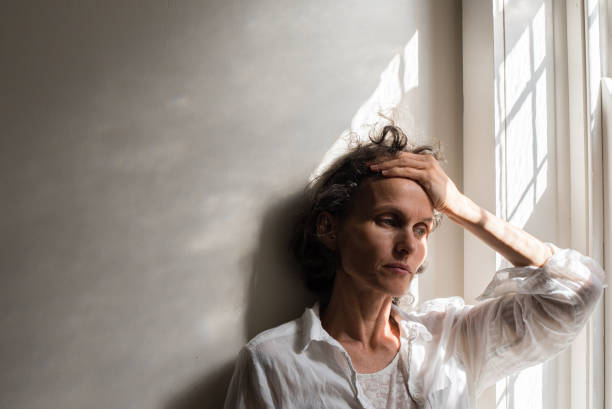 thoughtful tensed woman against wall by window at home - hand in hair imagens e fotografias de stock