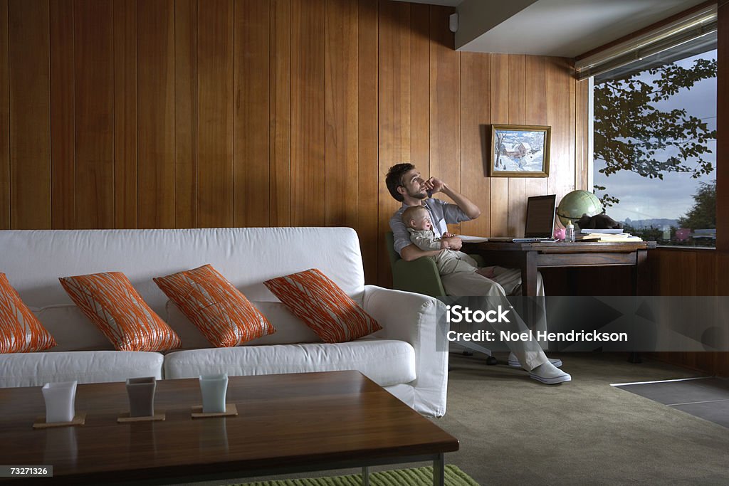 Father holding baby girl (6-9 months) and using cell phone in living room  20-29 Years Stock Photo