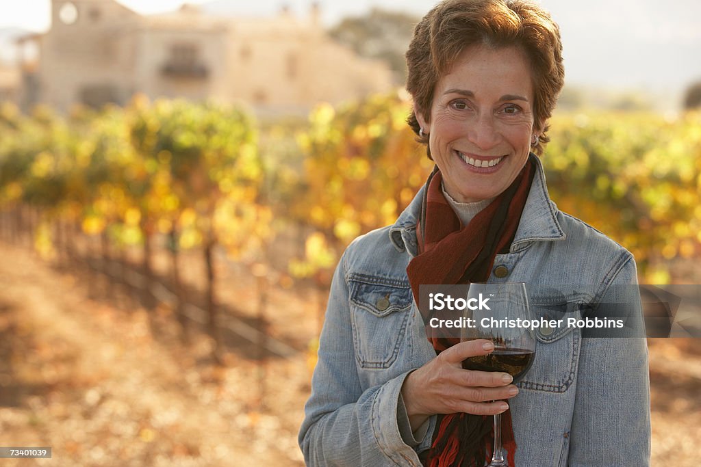 Donna matura in piedi in vineyard, che tiene un bicchiere di vino, smilin - Foto stock royalty-free di Azienda vinicola
