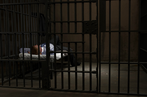 African American prisoner in orange uniform sits on the bed, reads Bible in prison cell. Prison officer walks, watches criminal. Inmate serves imprisonment term for crime in jail or detention center.