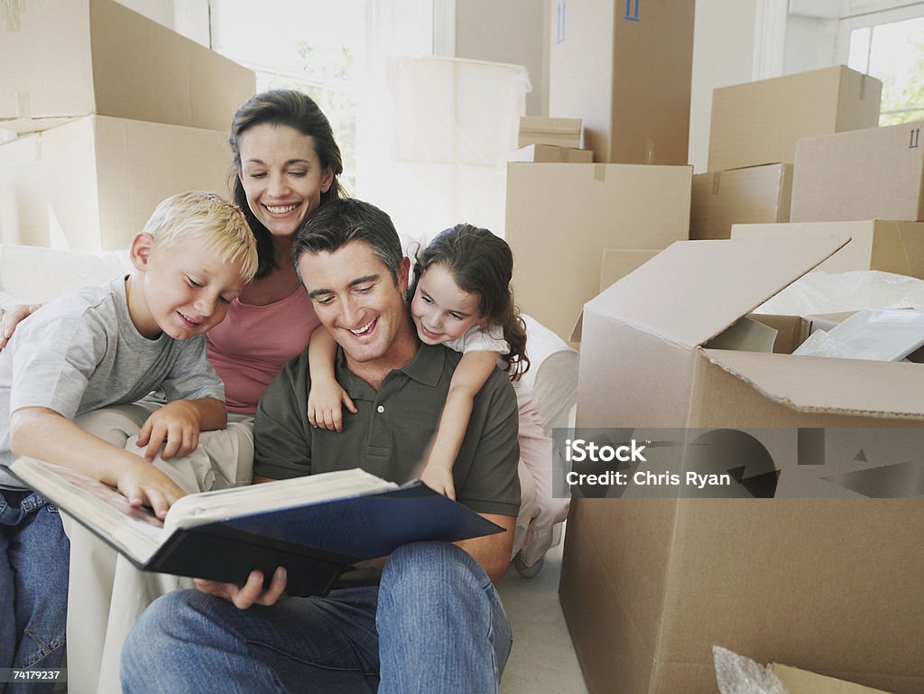 Man and woman with boy and girl looking at photo album in house with cardboard boxes  Photo Album Stock Photo