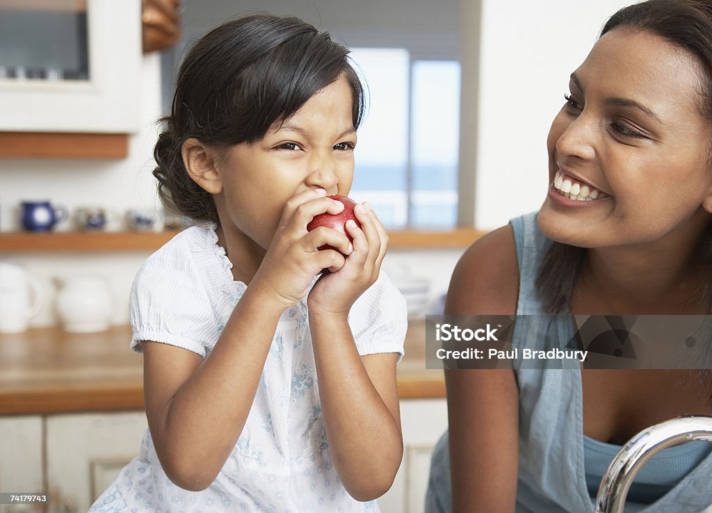 Mãe na cozinha com filha comendo maçã vermelha - Foto de stock de Comer royalty-free