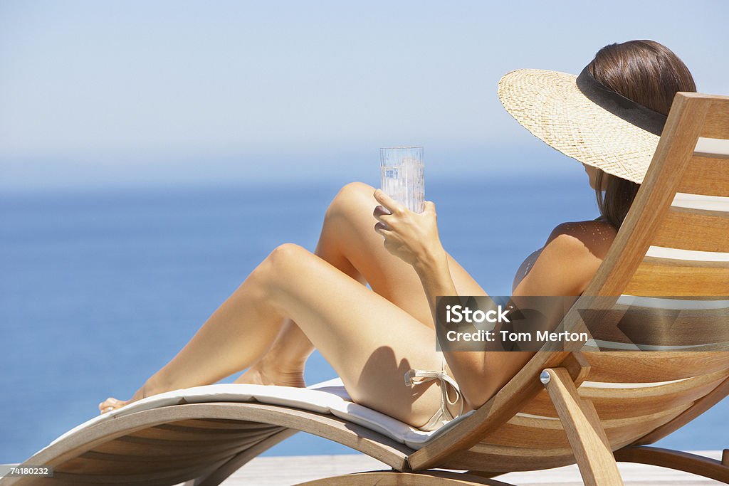 Mulher de chapéu de palha banhos de sol em uma cadeira de madeira com bebidas - Foto de stock de Só Uma Mulher royalty-free