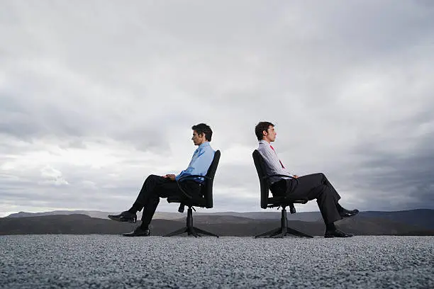 Photo of Two men sitting in office chairs outdoors with their backs against one another