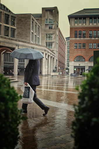 Paternoster Square outside St Paul's Cathedral in London, UK with rain falling heavily and a single businessman with an umbrella walking across the place.