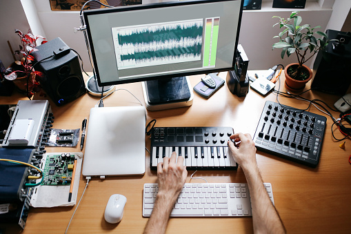 Overhead image of a young woman working in his home made apartment studio. Composing music, writing texts, doing a freelance job. Close up image, showing different hardware equipment, writing and music keyboards connected to different computer units.