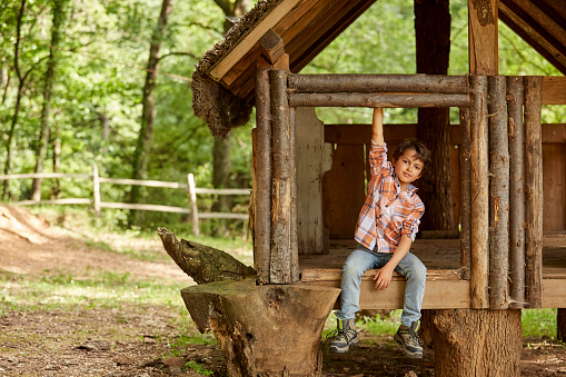 Smiling boy sitting in tree house. Full length of male enjoying in forest. He is wearing casuals.