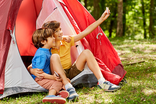 Happy brothers are taking selfie by tent. Smiling boys are camping in forest. They are spending leisure time.