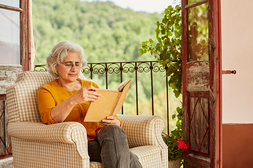 Senior woman reading book. Female is sitting on chair against balcony. She is wearing casuals.
