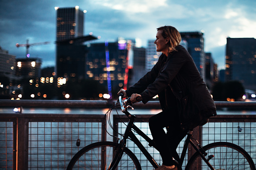 An adult woman rides her bicycle on the waterfront Eastside Esplanade in Portland, Oregon, a smile on her face as she explores the city night life.