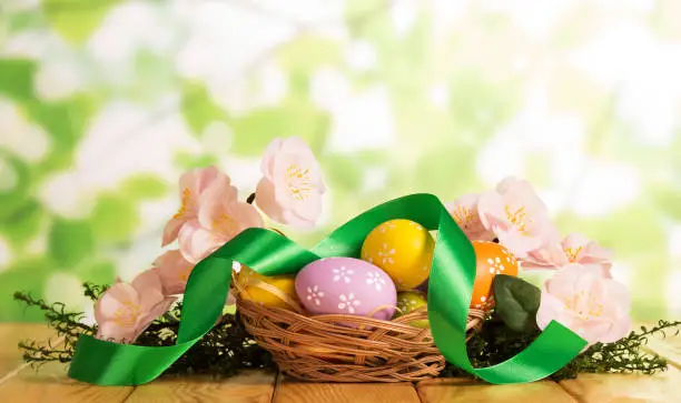Colorful Easter eggs in the basket, grass, branch with flowers and ribbon on abstract green background