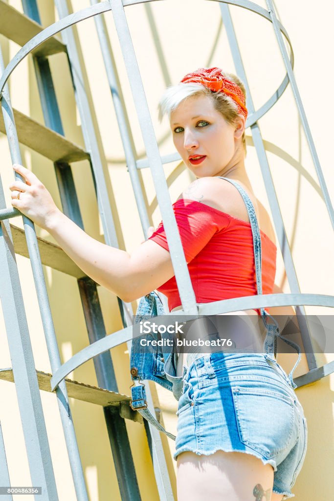 Jovem garota Pin-up loira com um laço vermelho em uma escada de emergência. Mulher Pin-up do conceito. - Foto de stock de Adulto royalty-free