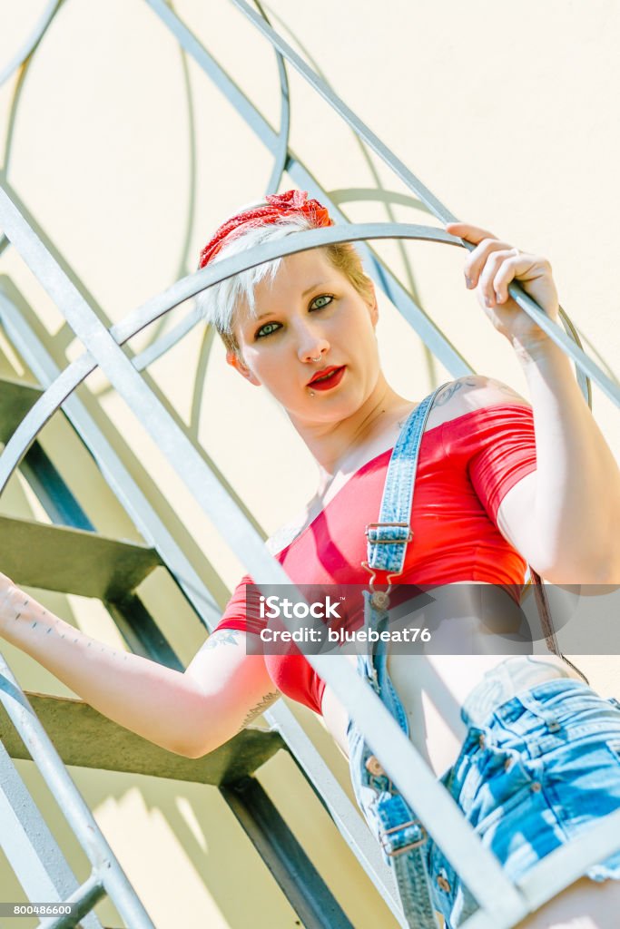 Jovem garota Pin-up loira com um laço vermelho em uma escada de emergência. Mulher Pin-up do conceito. - Foto de stock de Adulto royalty-free
