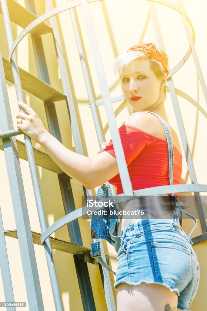 Jovem garota Pin-up loira com um laço vermelho em uma escada de emergência. Mulher Pin-up do conceito. - Foto de stock de Adulto royalty-free