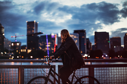 An adult woman rides her bicycle on the waterfront Eastside Esplanade in Portland, Oregon, a smile on her face as she explores the city night life.