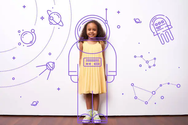 Cheerful cute African girl dreaming to be cosmonaut, she standing in exposure suit outer space drawing on white wall